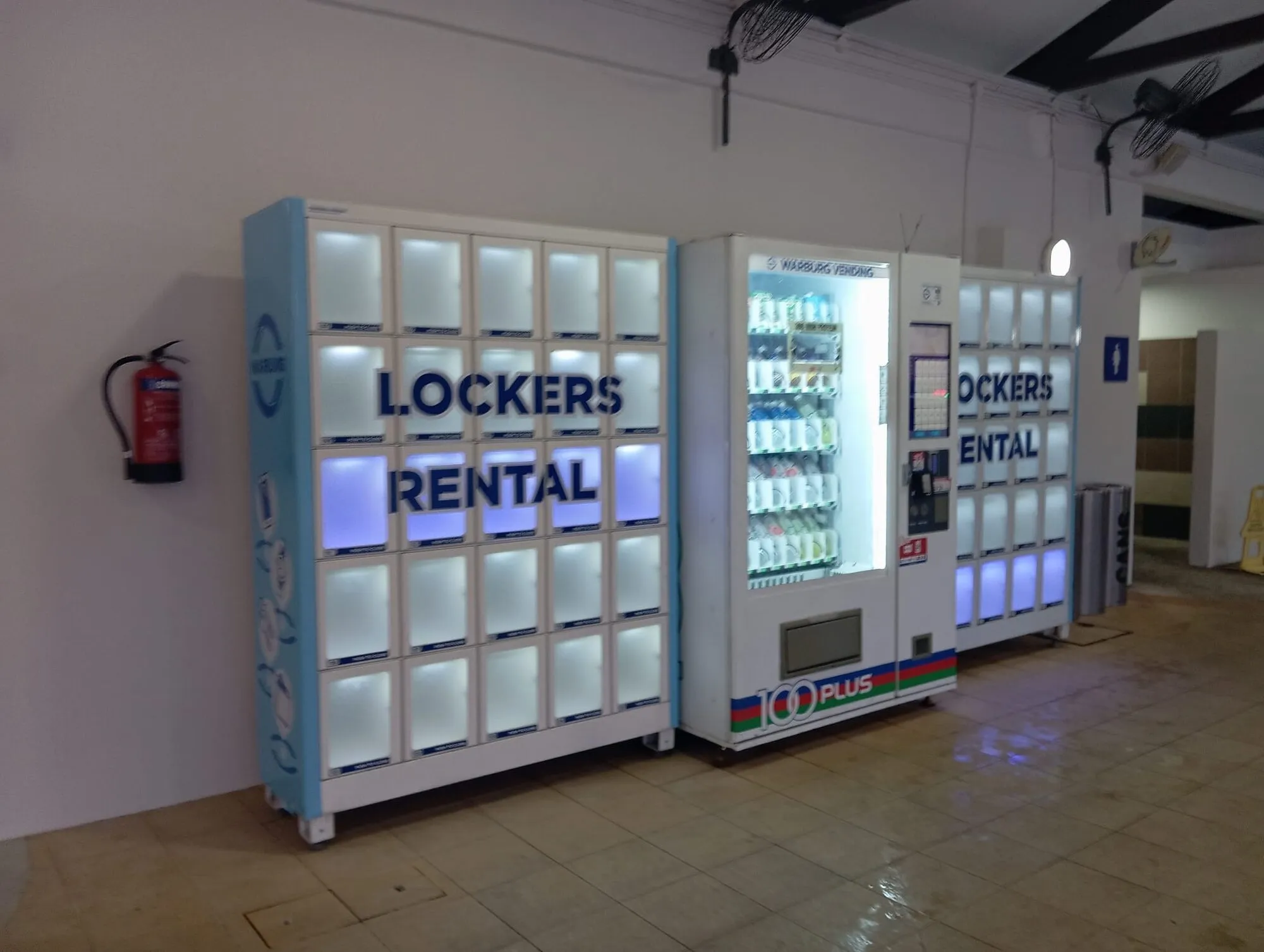 Lit locker rental machines and a 100PLUS drinks vending machine against a white wall